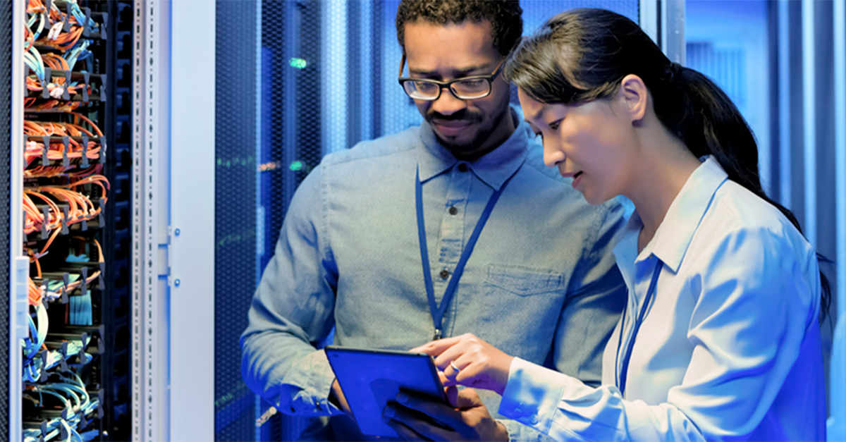 Two people working in a server room, reviewing information on a tablet near network racks with cables