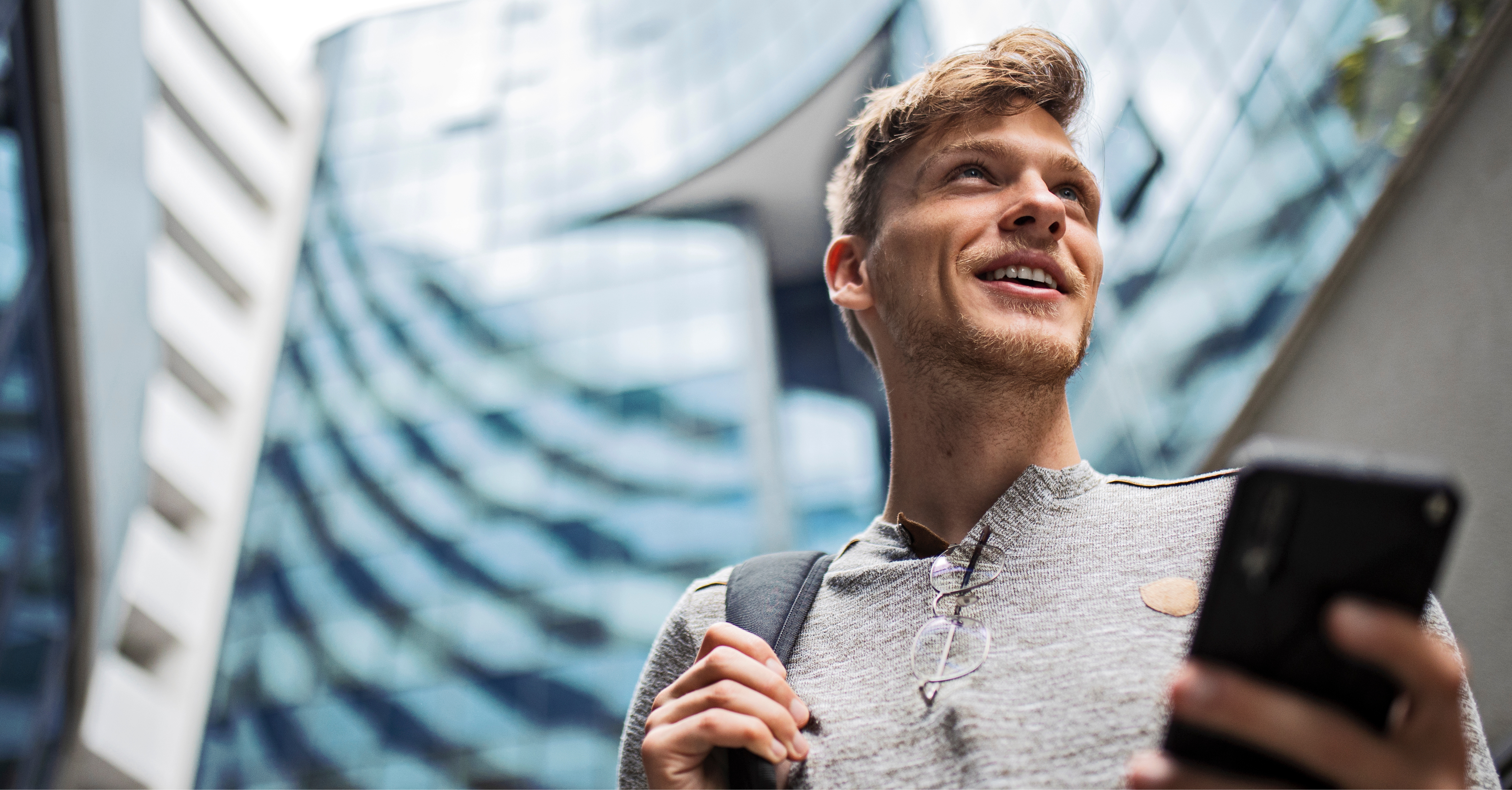 Person holding a smartphone and wearing a backpack, standing in front of modern glass buildings