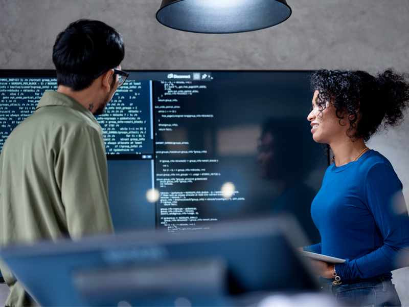 Two people stand in front of a large screen displaying code, discussing software development